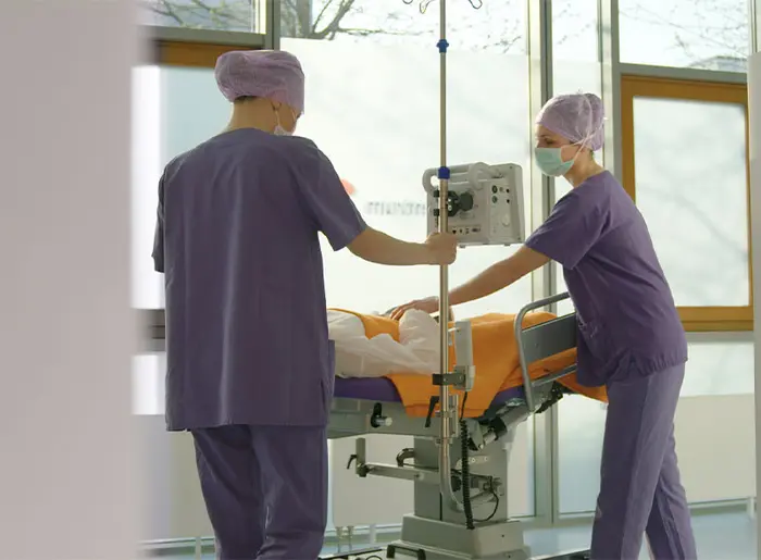 Zwei medizinische Assistentinnen in Operationskleidung bewegen einen Patientenwagen mit angeschlossenem Narkosemonitor.