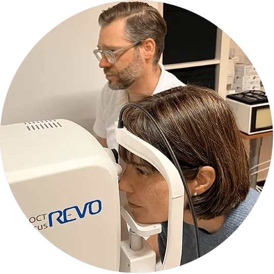 Patientin mit dem Kinn an der Stütze eines OCT REVO-Geräts, während der Augenoptikmeister am Steuerknüppel die 3D-Netzhautvermessung durchführt.