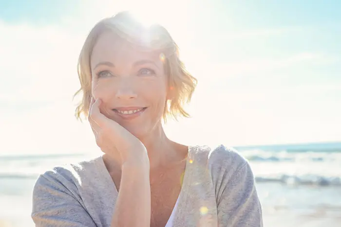 Eine Frau lächelt entspannt am Strand im Gegenlicht – Symbolbild für die positive Stimmung nach einer erfolgreichen Lidstraffung.